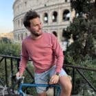 Model wearing The Climber Sweater by Classycles, standing with a vintage road bike against a Rome backdrop, featuring an embroidered cyclist design.